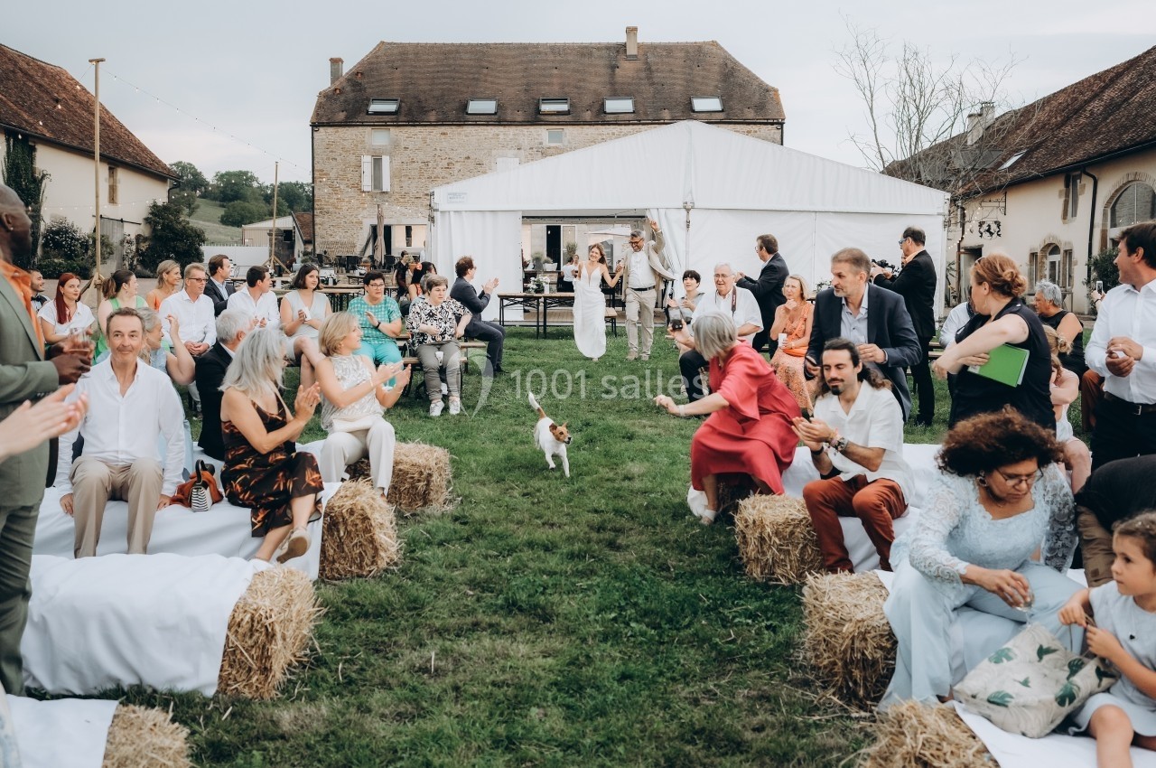 Des invités assis sur des bottes de paille assistent à une cérémonie en plein air devant une tente et une maison en pierre.