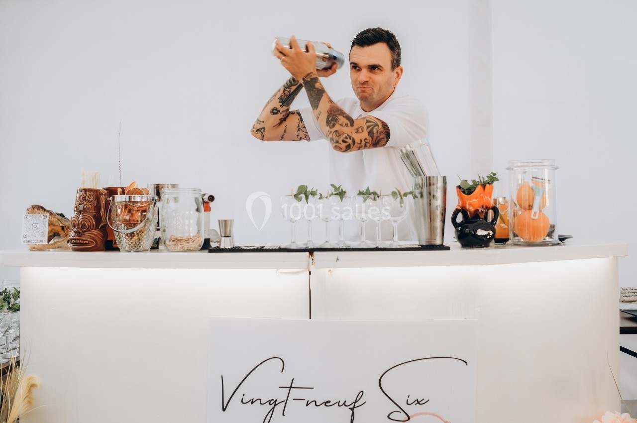 Un barman tatoué prépare un cocktail en secouant un shaker derrière un comptoir blanc décoré de verres et d'ingrédients.