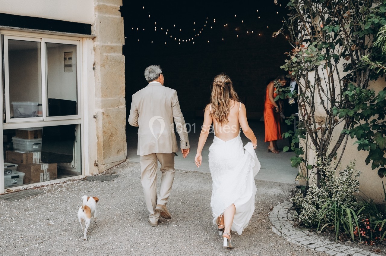 Une mariée en robe blanche marche avec un homme en costume beige et un petit chien vers une entrée décorée de guirlandes…