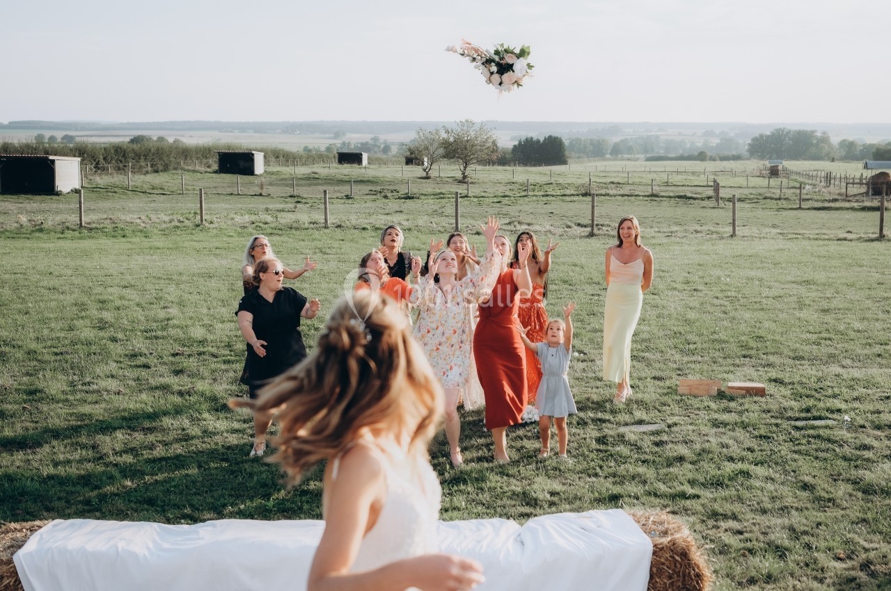 Une mariée lance son bouquet à un groupe de femmes dans un champ lors d'une réception en plein air.