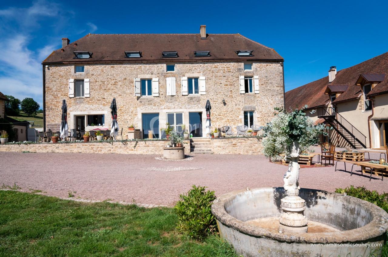 Façade d'une grande maison en pierre avec terrasse, parasols et une fontaine au premier plan, sous un ciel bleu.