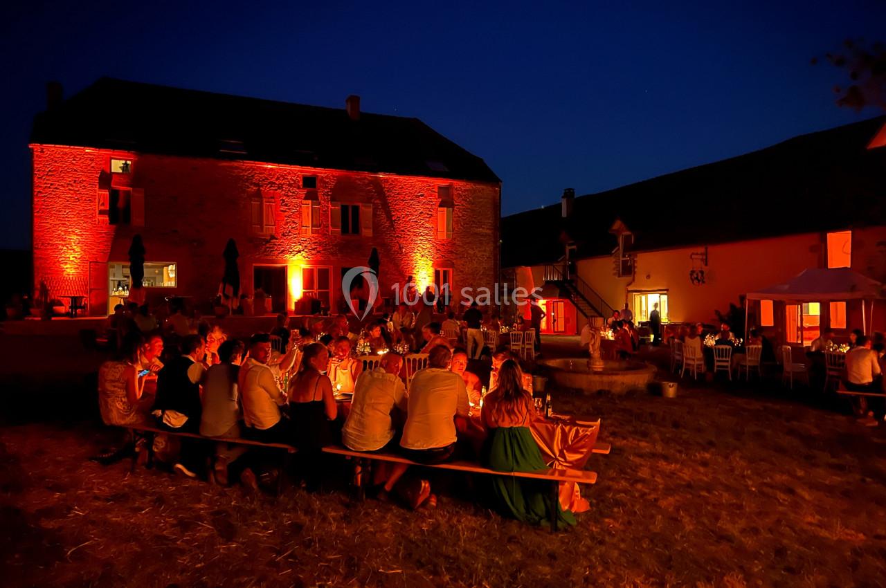 Groupe de personnes dînant en plein air devant un bâtiment en pierre illuminé de lumières rouges, de nuit.