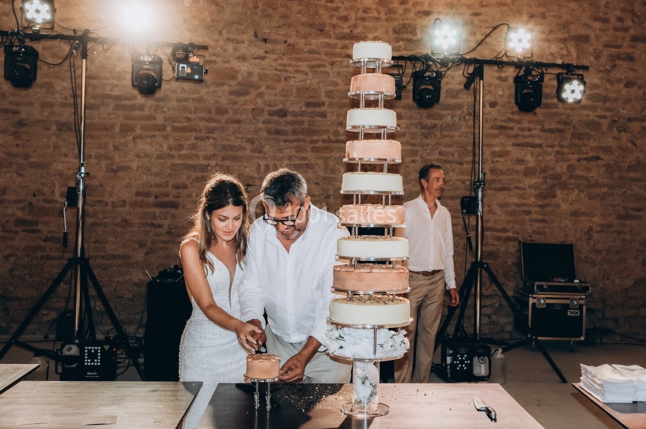 Un couple coupe une pièce montée lors d'une célébration, avec un mur en pierre et des éclairages en arrière-plan.
