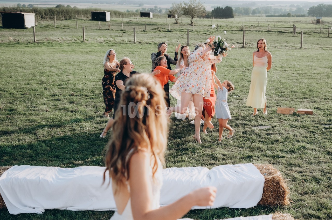 Une femme lance un bouquet de fleurs à un groupe de personnes dans un champ lors d'une célébration en plein air.