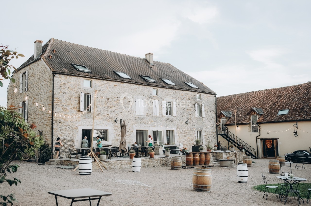 Grande maison en pierre avec volets blancs, terrasse animée, tonneaux décoratifs et ciel dégagé en arrière-plan.