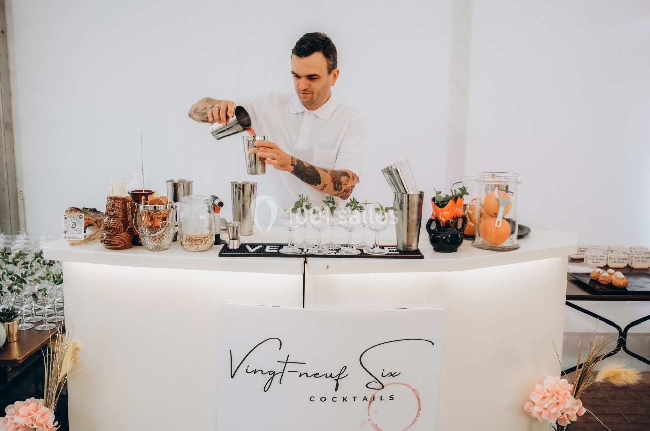 Un barman prépare un cocktail derrière un comptoir blanc décoré avec des ingrédients et des accessoires.