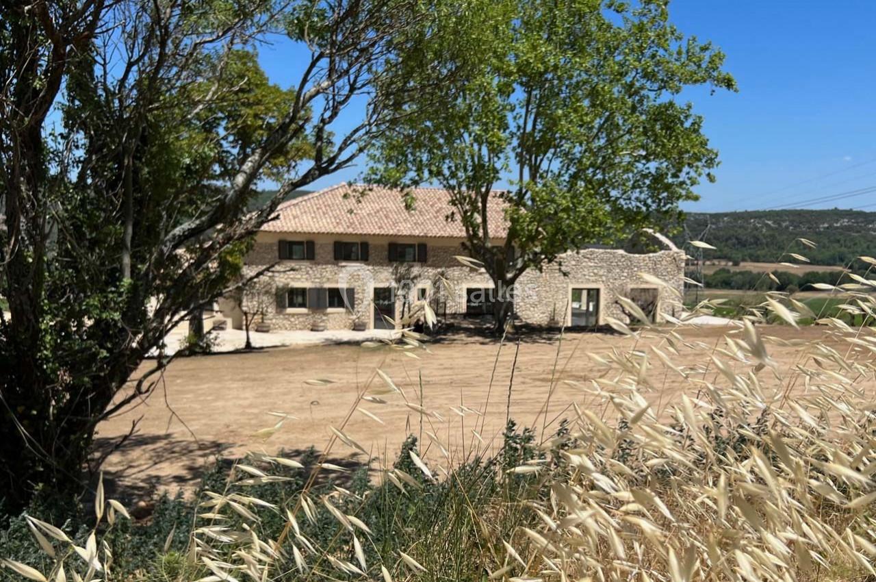 Maison en pierre entourée de végétation, avec un grand arbre à l'avant, sous un ciel bleu clair.