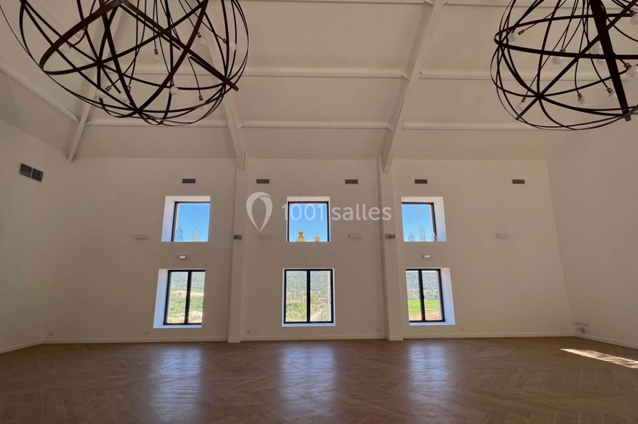 Grande salle lumineuse avec parquet en bois clair, hauts plafonds blancs et grandes fenêtres offrant une vue sur l'extérieur.