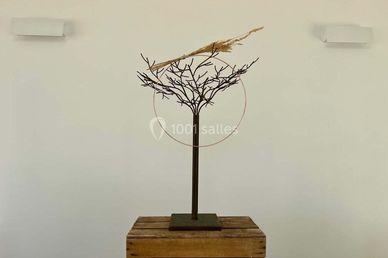 Sculpture minimaliste représentant un arbre en métal avec une tige végétale et un cercle, posée sur un socle en bois.
