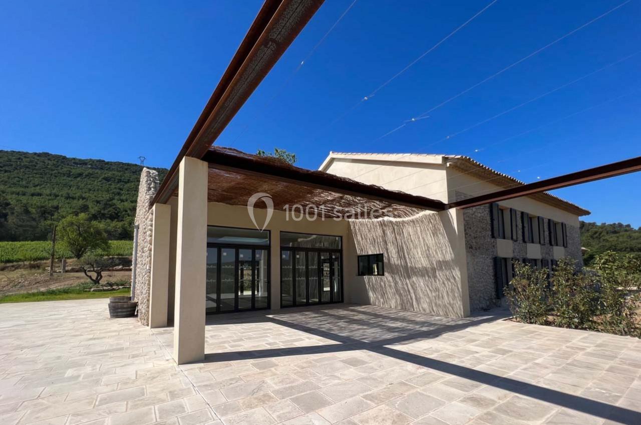 Façade d'un bâtiment moderne en pierre avec une terrasse ombragée, entouré de végétation et d'un ciel dégagé.