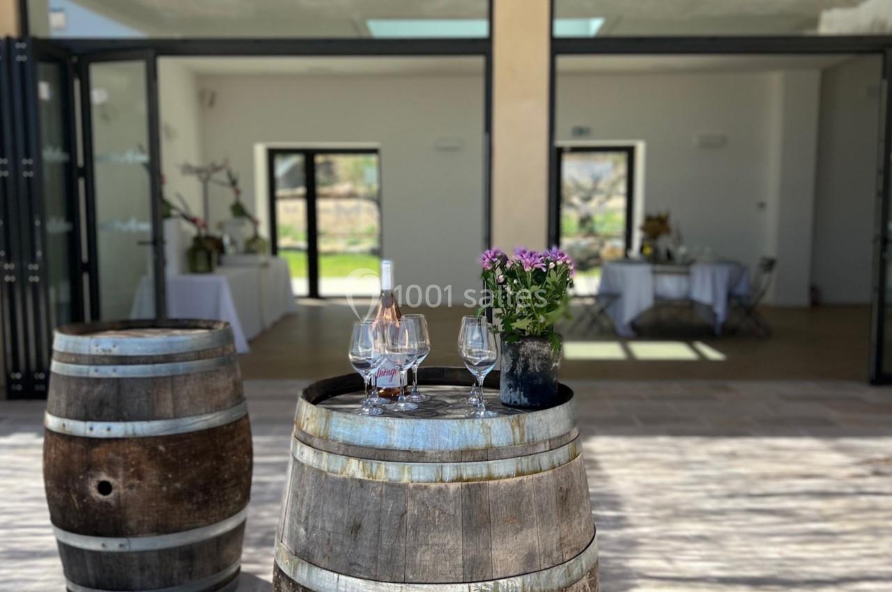 Deux tonneaux en bois servent de table avec une bouteille de vin rosé, des verres et des fleurs, devant une salle ouverte.