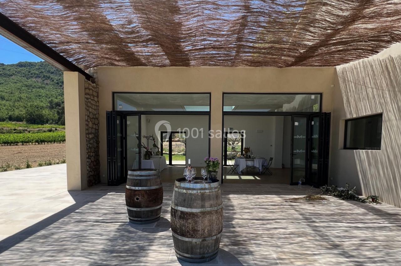 Terrasse ombragée avec deux tonneaux en bois, vue sur une salle vitrée et un paysage de vignobles en arrière-plan.