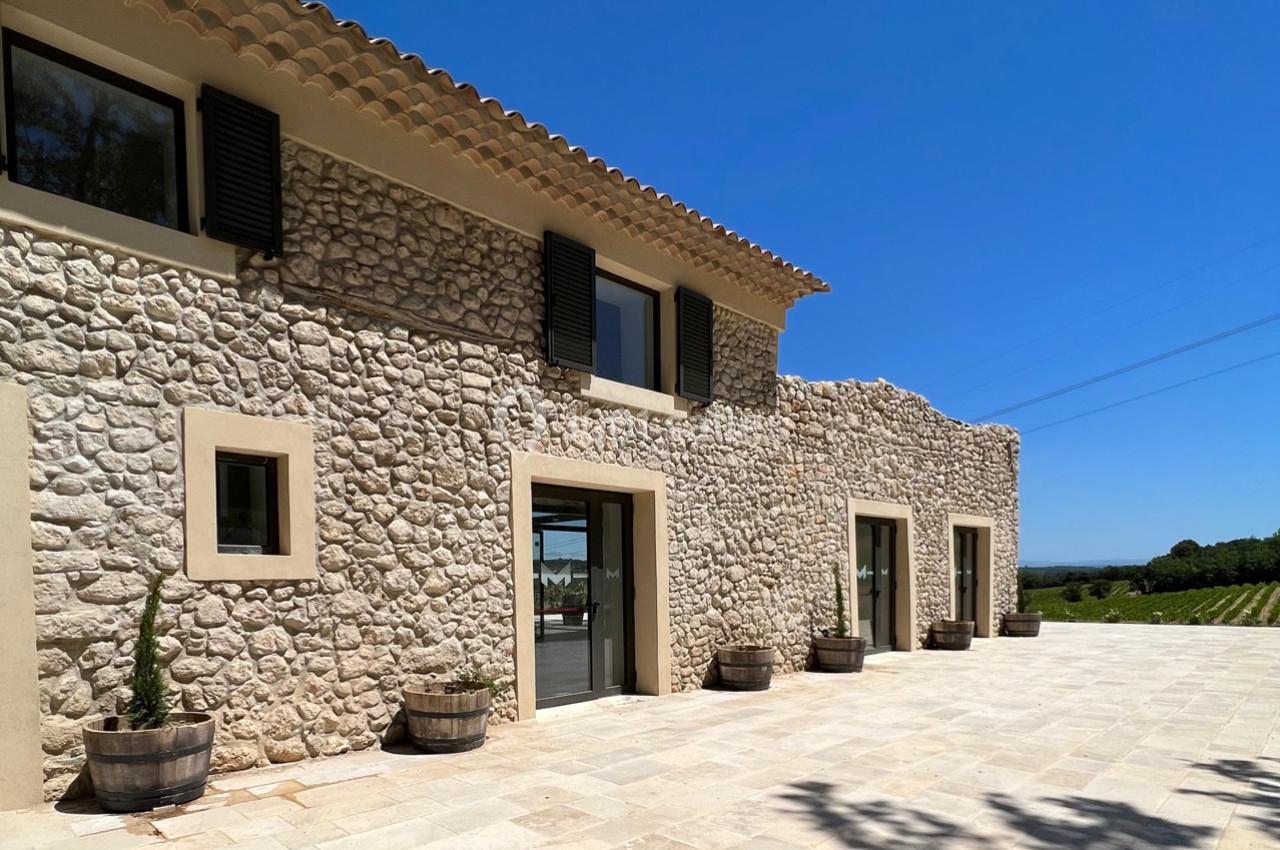 Façade en pierre d'une maison avec volets noirs, terrasse pavée et vue sur des vignes sous un ciel bleu.