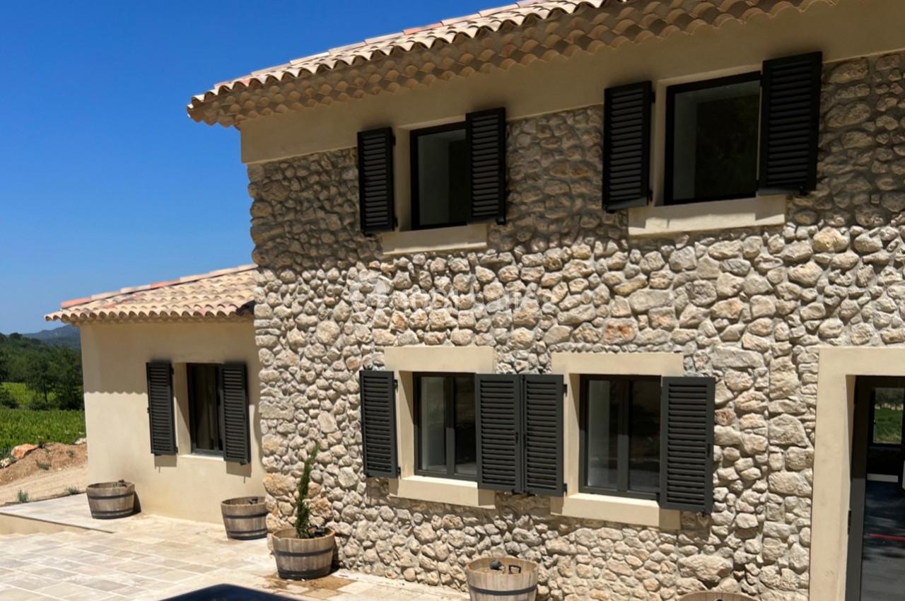 Façade en pierre d'une maison avec volets noirs, terrasse en pierre et barriques en bois, sous un ciel bleu.