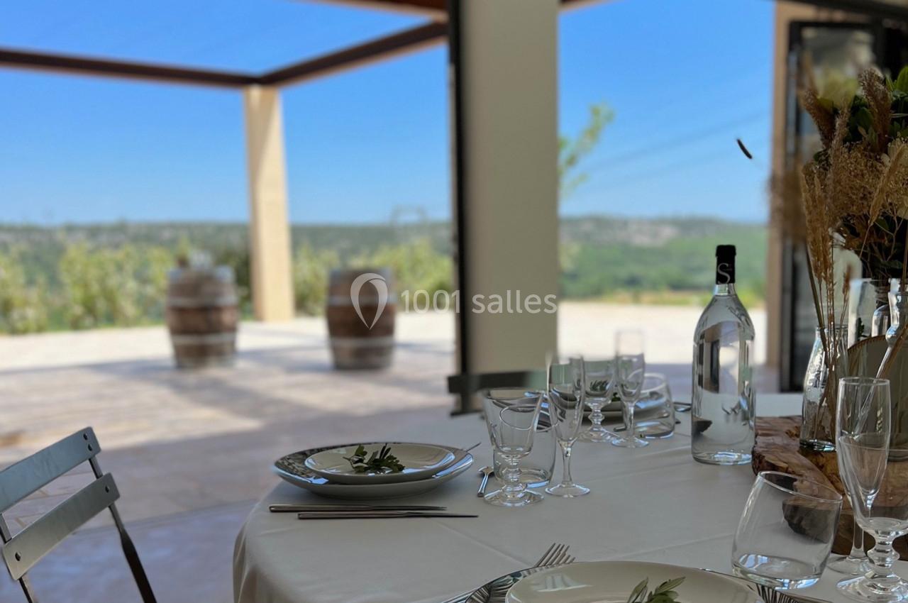 Table dressée avec vaisselle élégante, vue sur une terrasse ensoleillée et des barriques en arrière-plan.