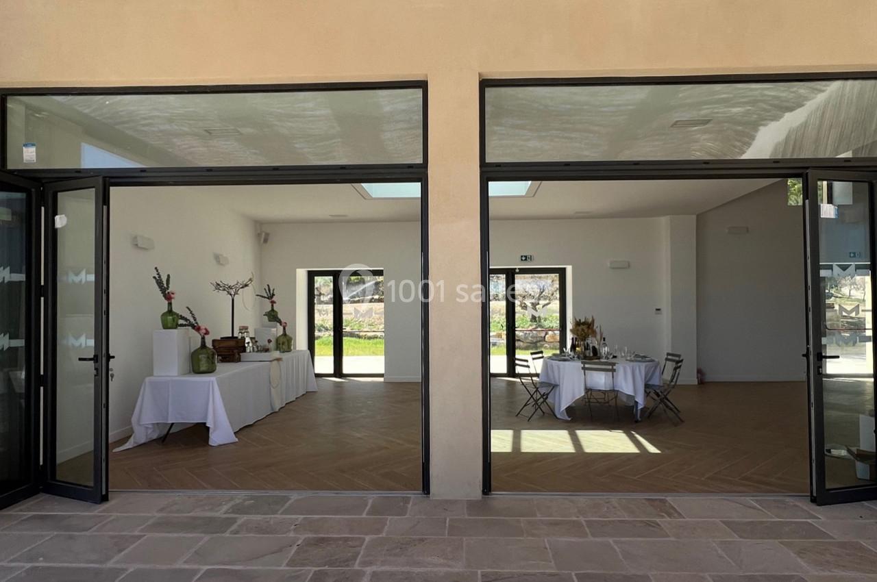 Salle lumineuse avec grandes baies vitrées, tables dressées et décorations minimalistes, vue sur un extérieur ensoleillé.