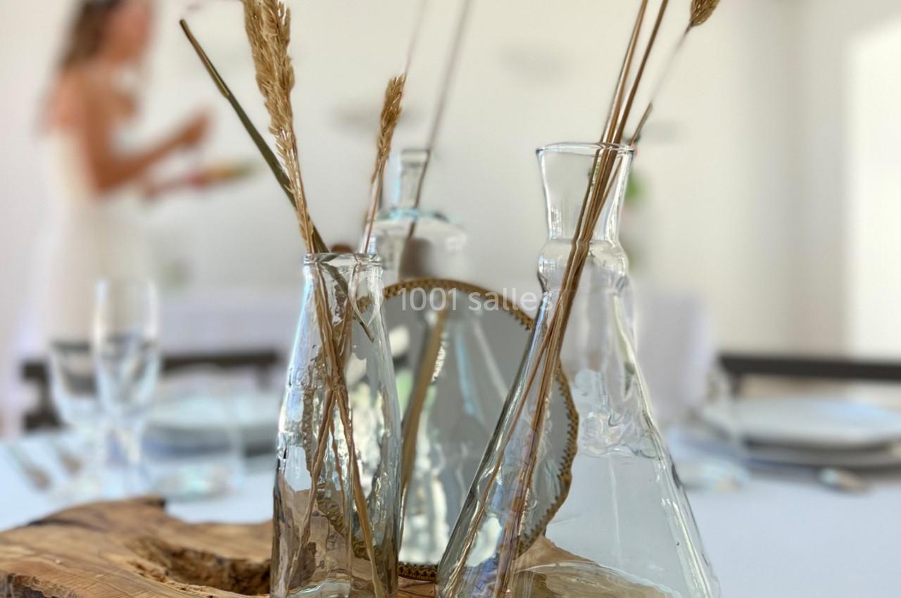 Vases en verre avec des herbes séchées sur une table décorée, en arrière-plan une silhouette floue.