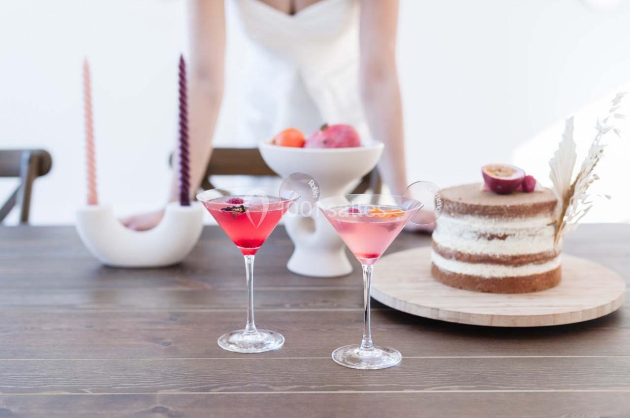 Deux cocktails roses sur une table en bois, accompagnés d'un gâteau, de fruits et de bougies décoratives.