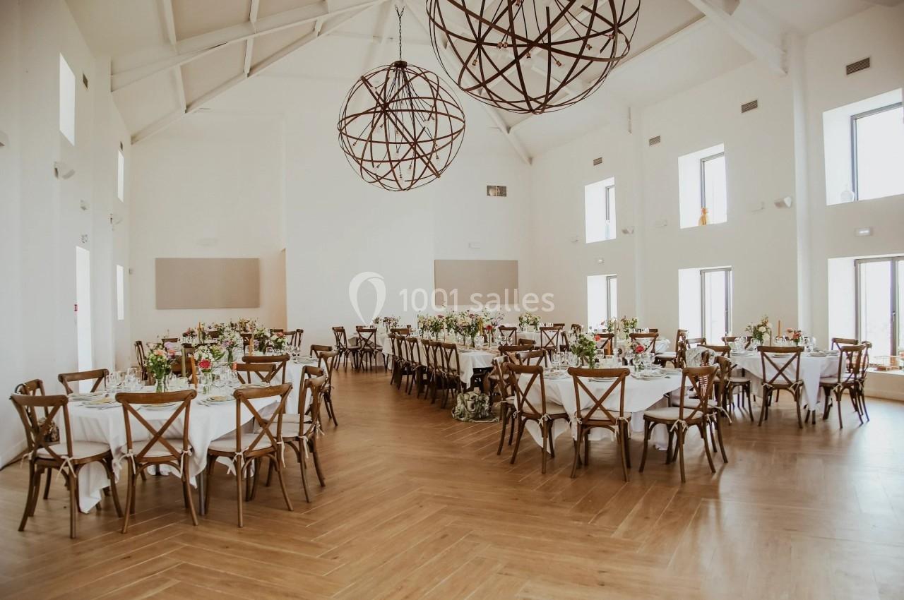 Salle lumineuse avec tables rondes dressées pour un événement, décorée de fleurs et éclairée par des suspensions métalliques.
