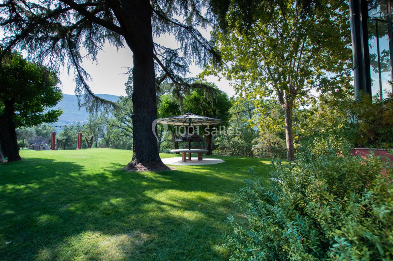 Espace vert avec pelouse, arbres et une table de pique-nique sous un abri, entouré de végétation et de collines en arrière…