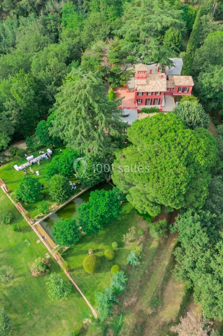 Vue aérienne d'une maison rouge entourée d'un grand jardin verdoyant avec arbres, pelouses et allées aménagées.