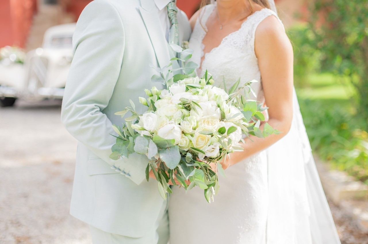 Un couple en tenue de mariage tient un bouquet de roses blanches et de feuillage, avec une voiture ancienne en arrière-plan.