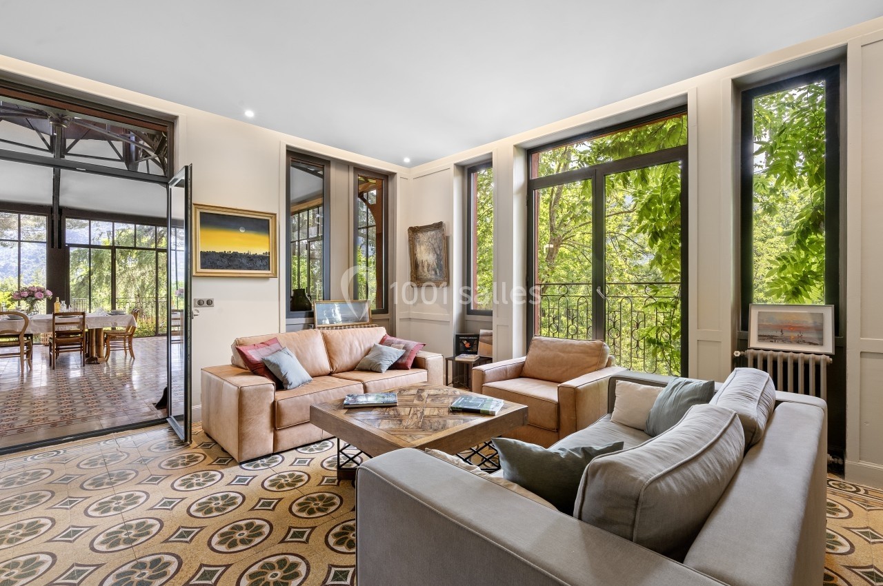Salon lumineux avec canapés, table basse et grandes fenêtres donnant sur un espace verdoyant.