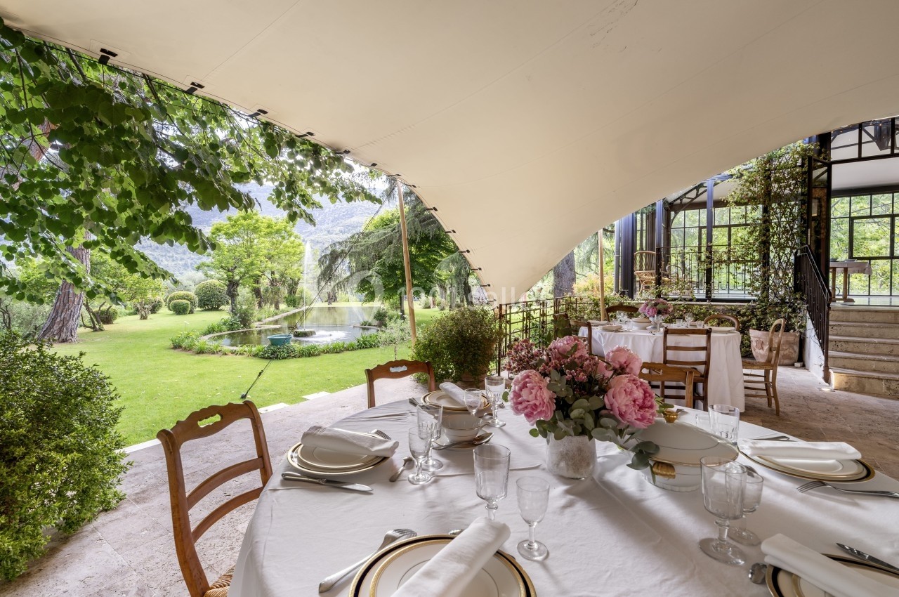 Table dressée avec vaisselle élégante sous une tente ouverte, offrant une vue sur un jardin verdoyant avec un étang.