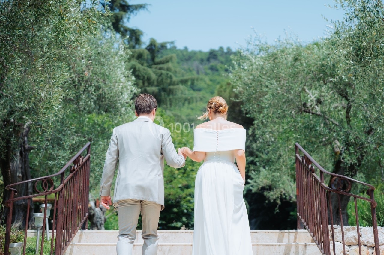 Un couple en tenue de mariage descend un escalier entouré d'arbres et de verdure sous un ciel clair.