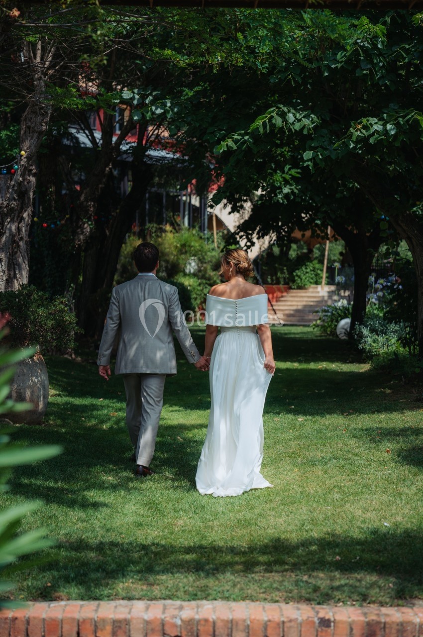 Un couple en tenue de mariage marche main dans la main dans un jardin verdoyant.