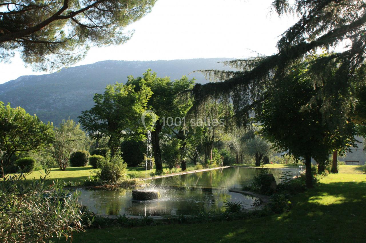 Étang circulaire avec une fontaine entouré de végétation luxuriante, sous un ciel clair et une montagne en arrière-plan.