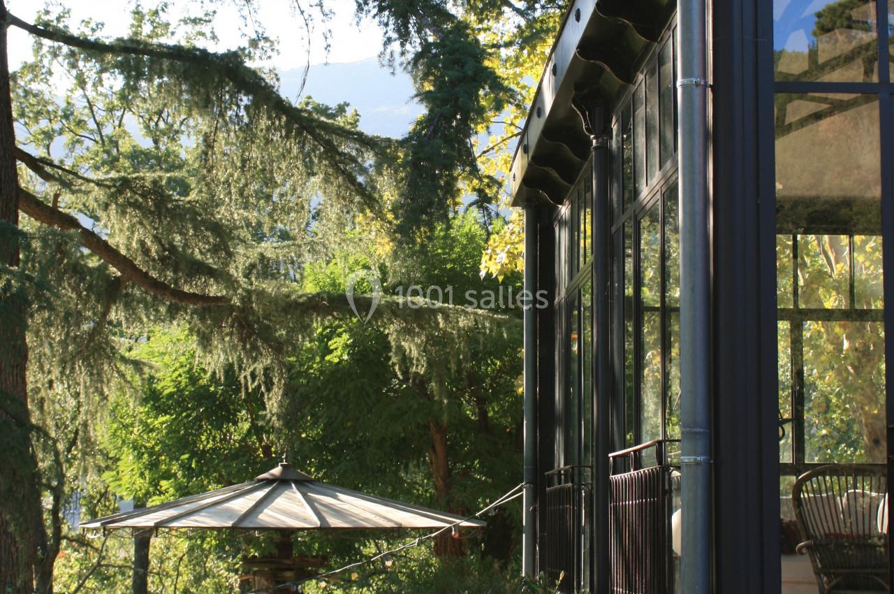 Véranda en verre et métal donnant sur un jardin arboré avec une table et un parasol sous les arbres.