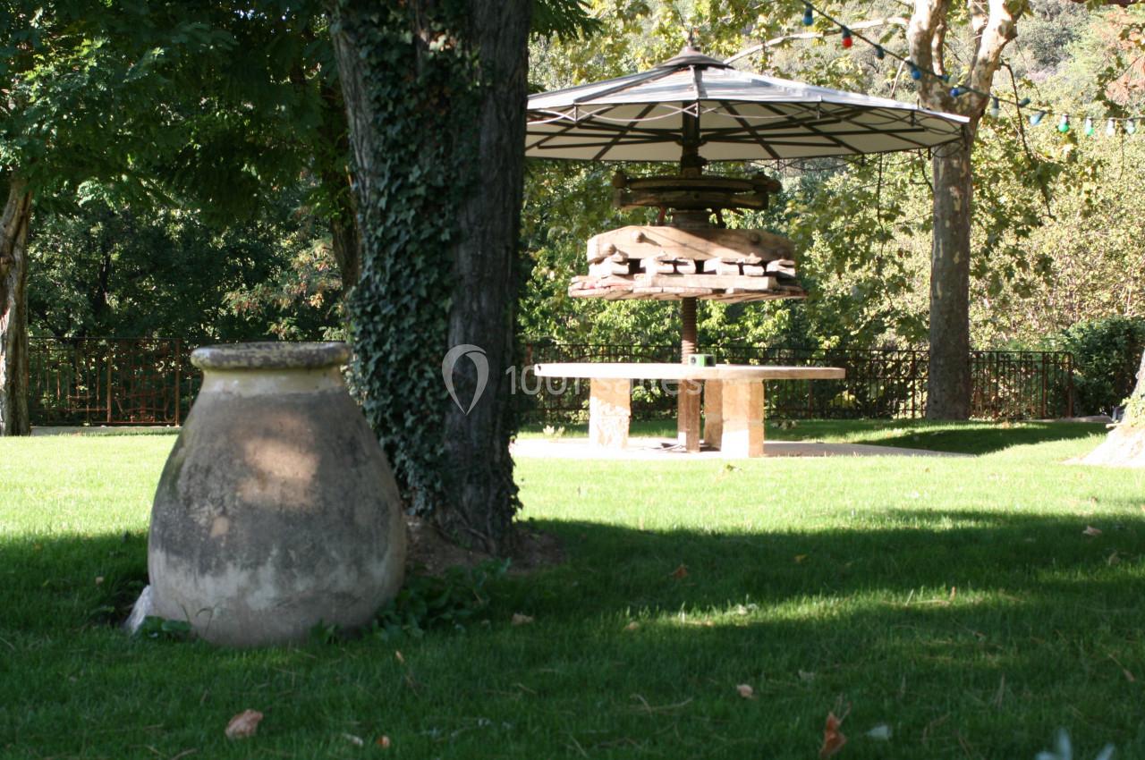 Un jardin verdoyant avec un grand arbre, une jarre en pierre et une table en pierre sous un parasol.