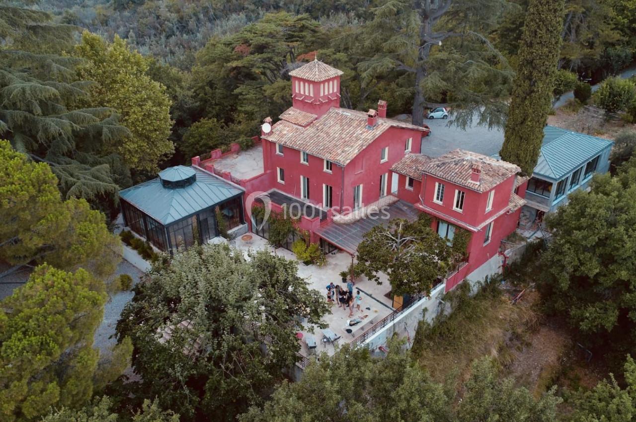 Vue aérienne d'une grande maison rouge entourée d'arbres, avec une terrasse et une verrière attenante.