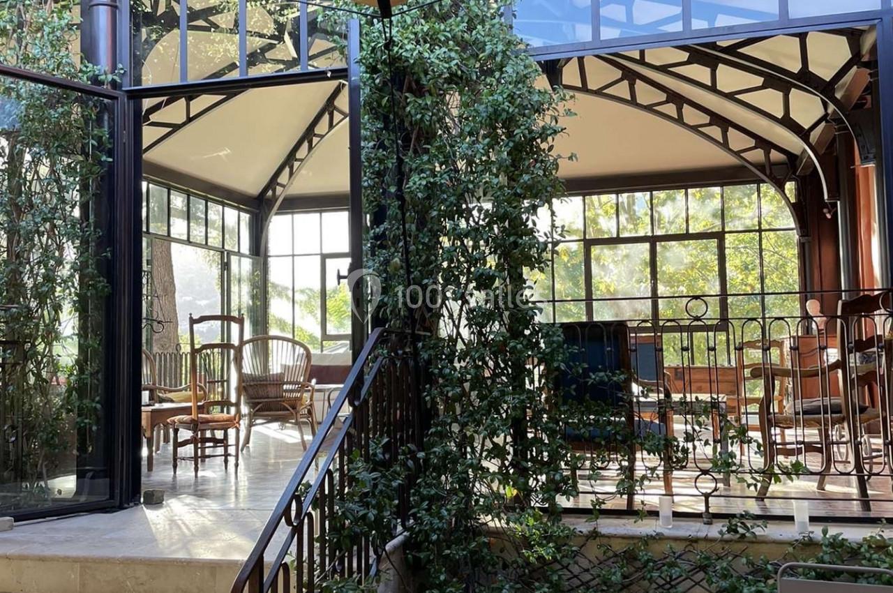 Intérieur lumineux avec verrière, plantes grimpantes, mobilier en bois et métal, et escalier menant à une terrasse.