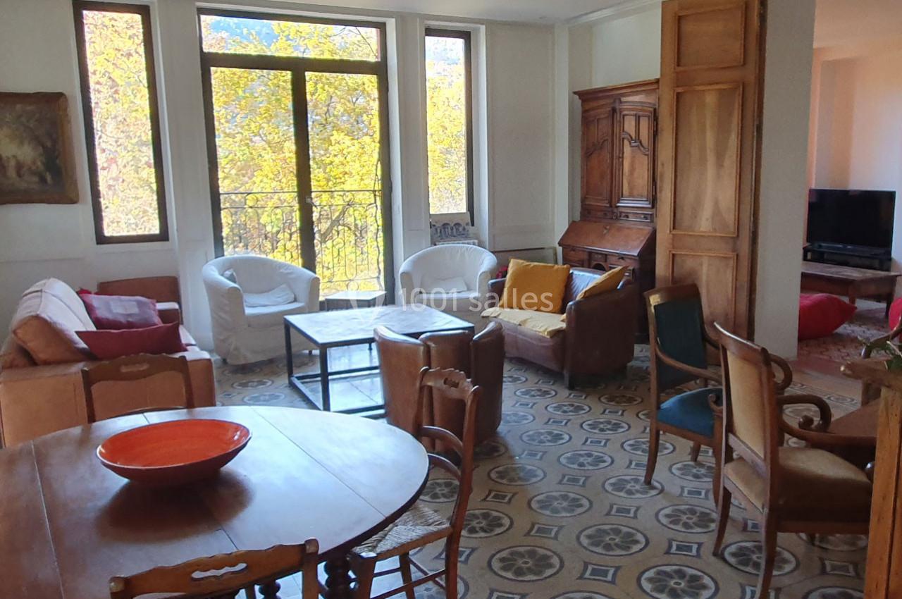 Salon lumineux avec grandes fenêtres, mobilier varié, table ronde en bois et sol en carreaux décoratifs.