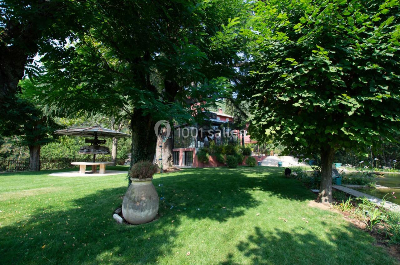 Jardin verdoyant avec pelouse, arbres, pot décoratif, table ombragée et bâtiment en arrière-plan.