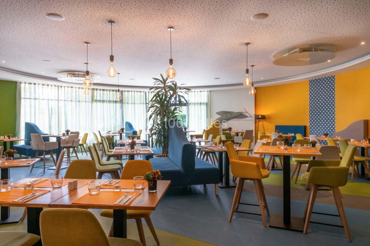 Salle de restaurant lumineuse avec tables en bois, chaises colorées et décoration moderne, éclairée par des suspensions.