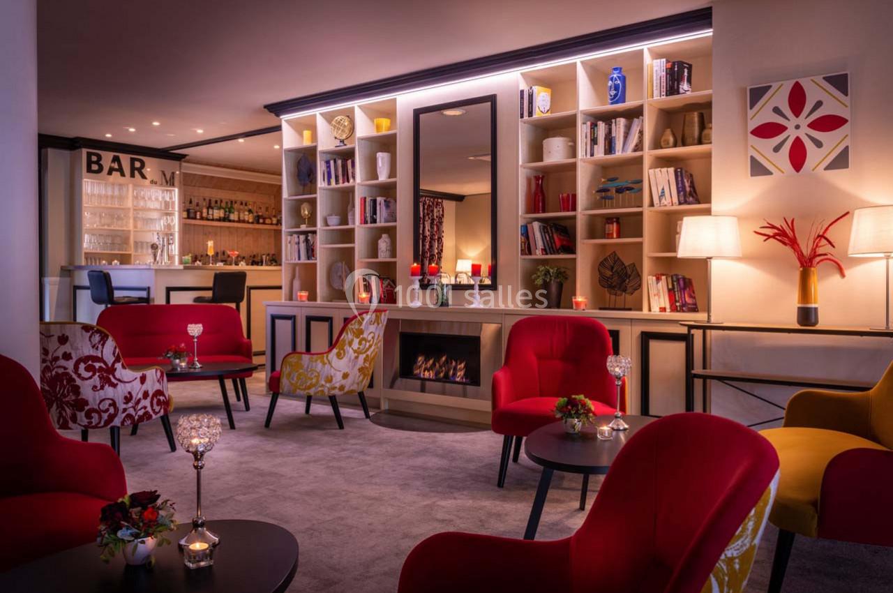 Salon chaleureux avec fauteuils colorés, cheminée centrale, étagères décoratives et bar en arrière-plan.