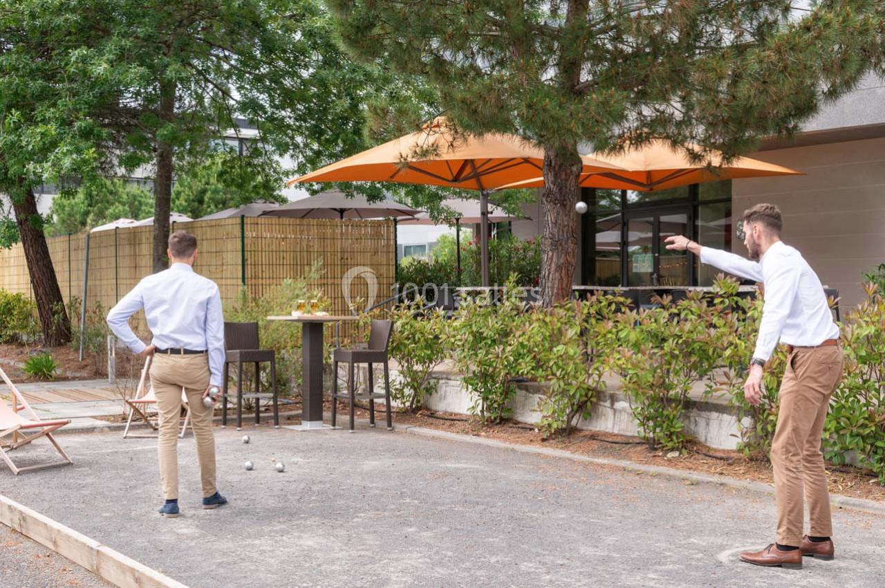 Deux hommes jouent à la pétanque sur un terrain extérieur entouré de végétation et de parasols.