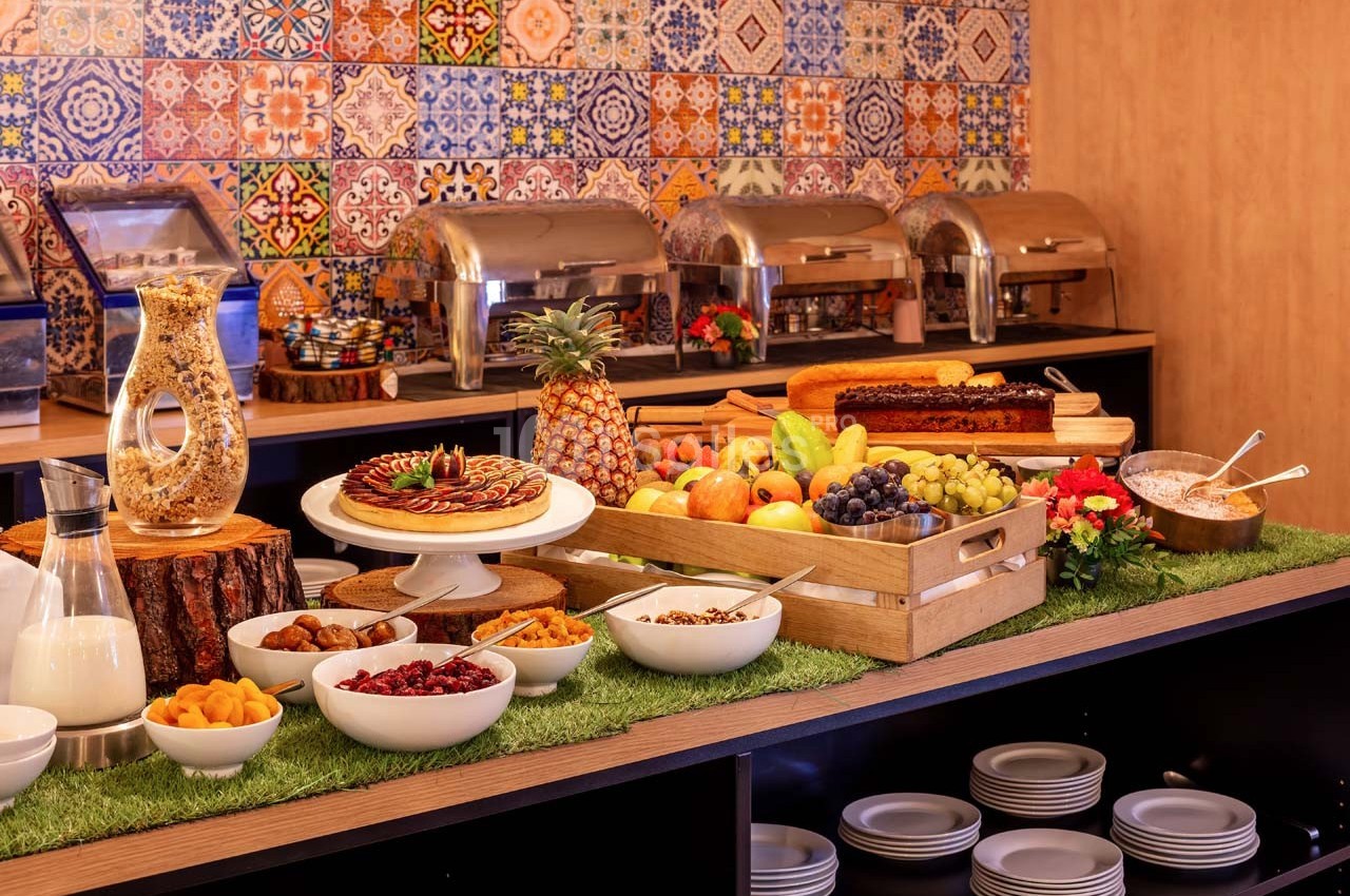 Buffet avec fruits frais, pâtisseries, céréales et plats chauds, présenté sur une table décorée devant un mur coloré.