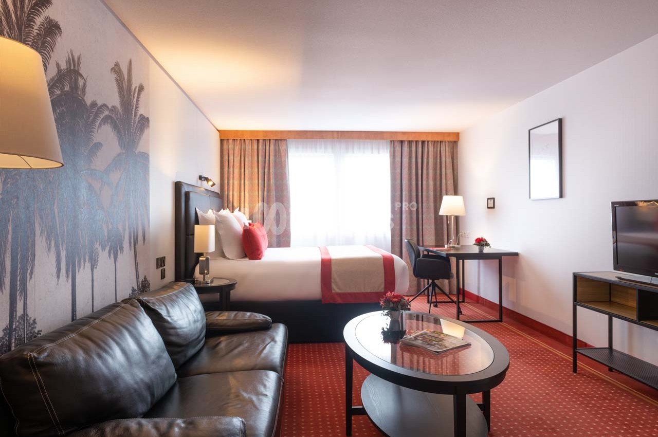 Chambre d'hôtel lumineuse avec lit double, canapé en cuir, bureau, téléviseur et décoration murale de palmiers.