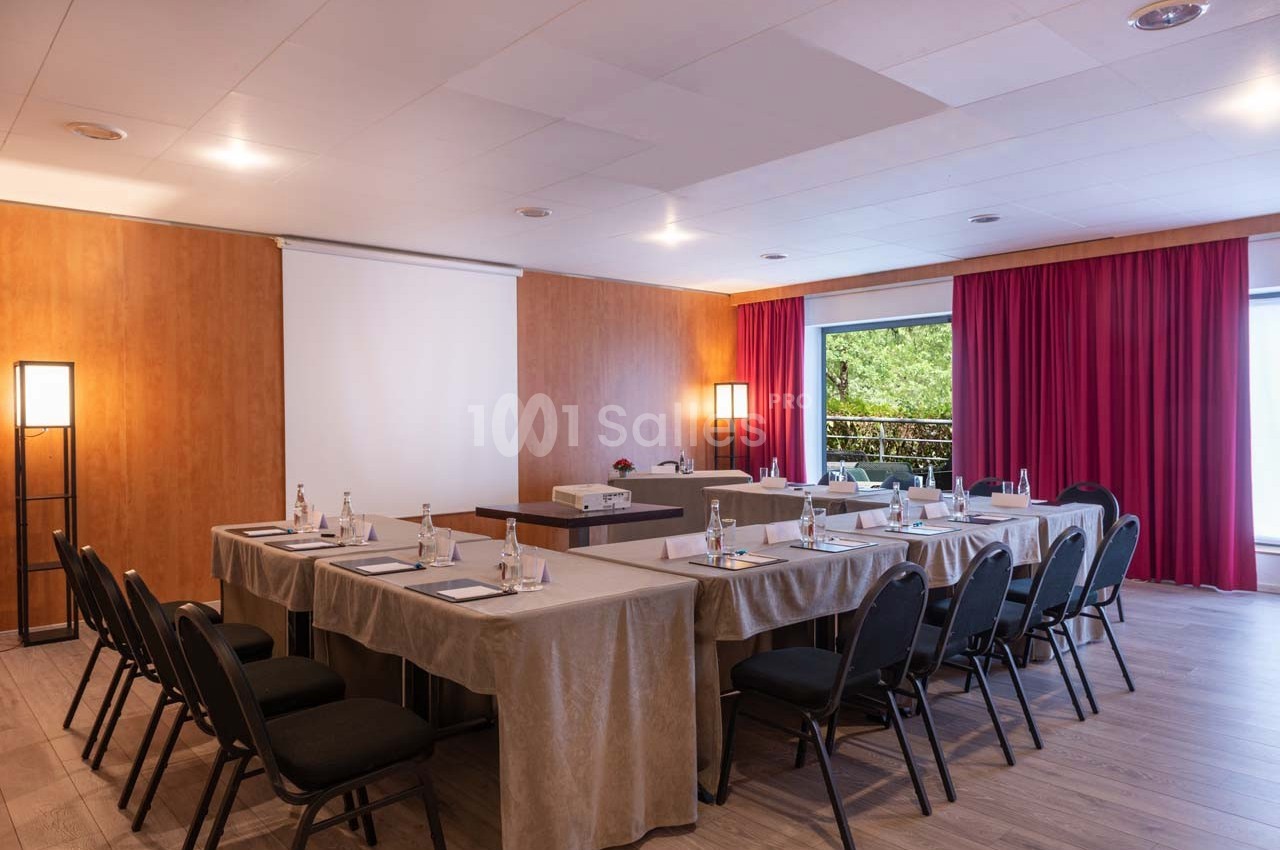 Salle de réunion lumineuse avec tables en U, chaises noires, projecteur, rideaux rouges et vue sur un espace extérieur…