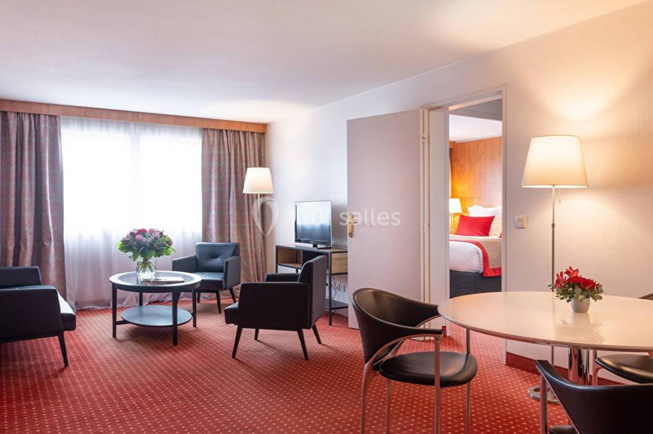 Salon d'hôtel avec fauteuils, table basse, table à manger, lampes et vue sur une chambre adjacente.