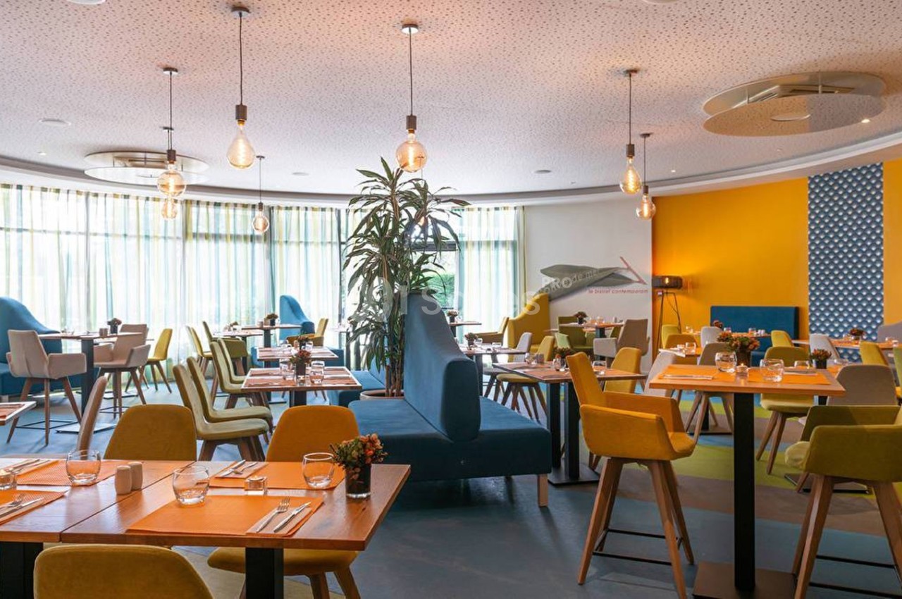 Salle de restaurant lumineuse avec tables dressées, chaises colorées et décoration moderne.