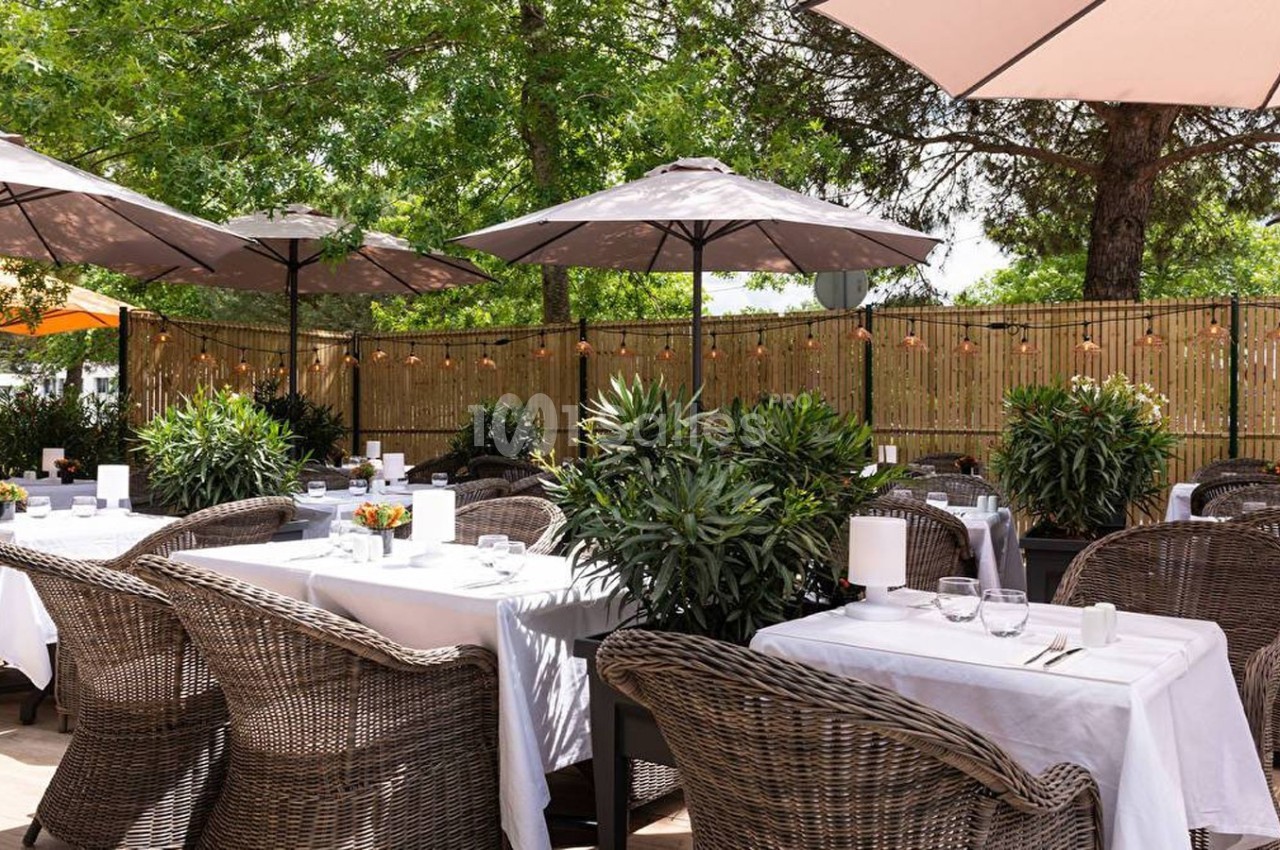 Terrasse de restaurant avec tables dressées, chaises en rotin, parasols et végétation environnante sous un ciel dégagé.