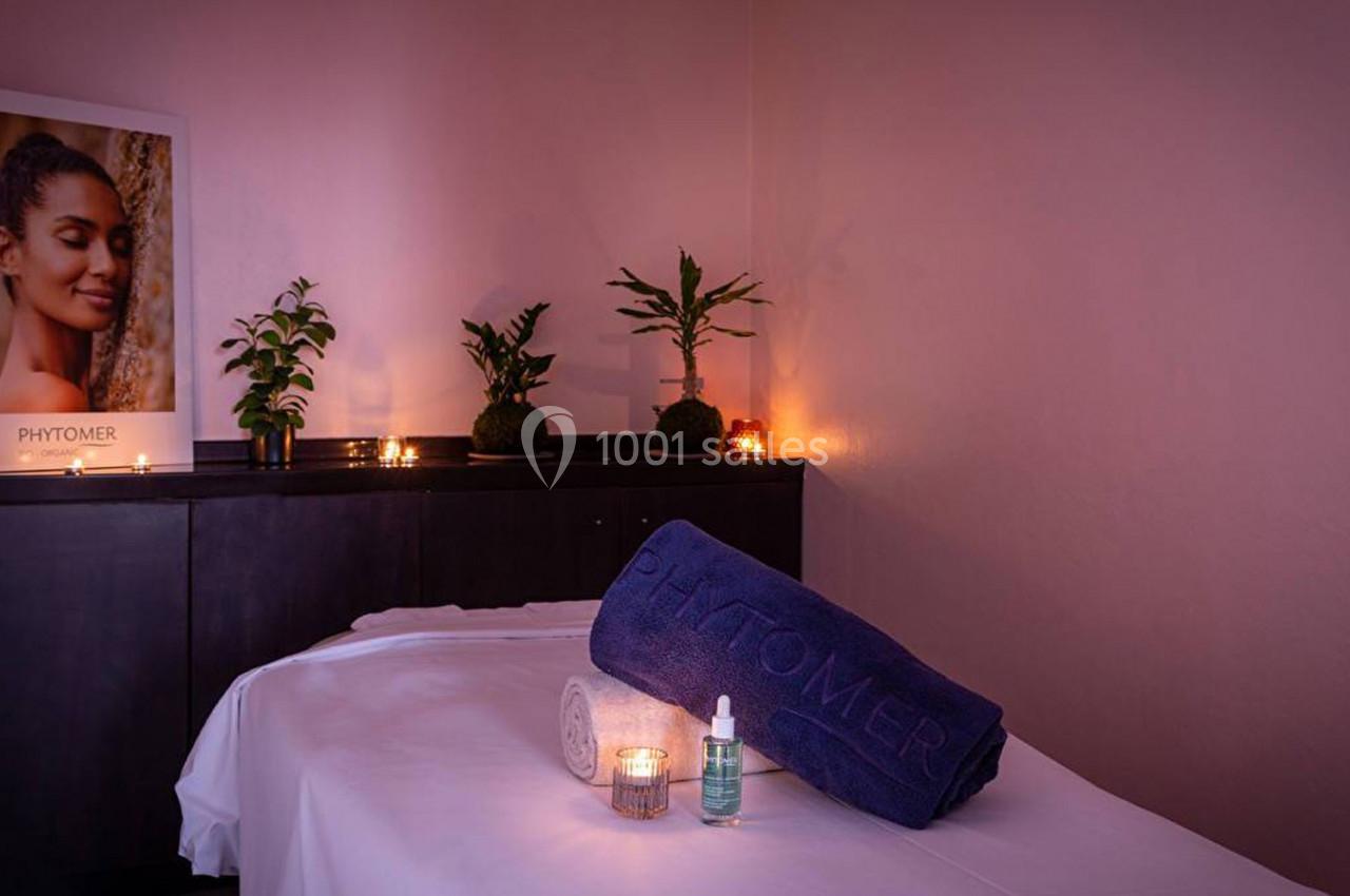 Salle de soins avec table de massage, serviette, produits de beauté, bougies allumées et plantes décoratives.