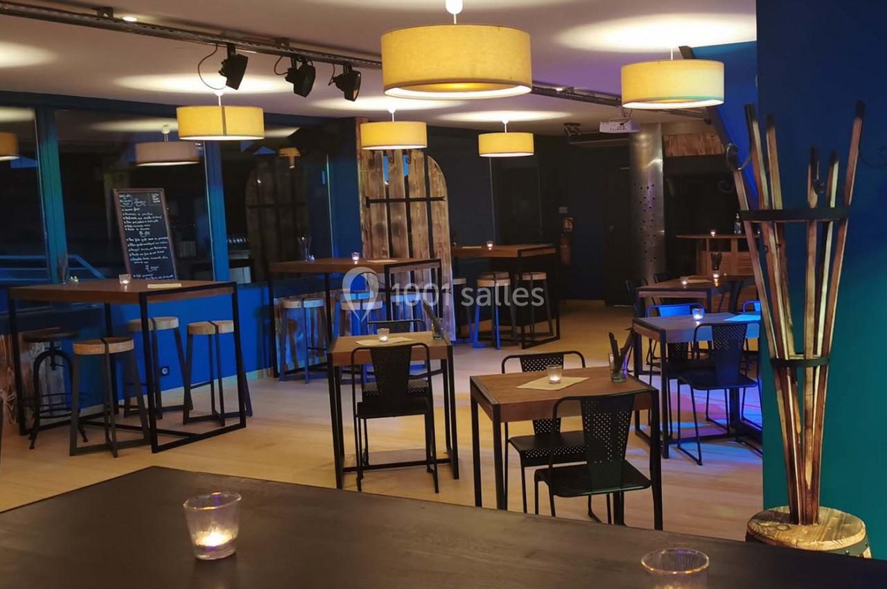 Salle moderne avec tables hautes et basses, éclairée par des suspensions lumineuses et des bougies sur les tables.
