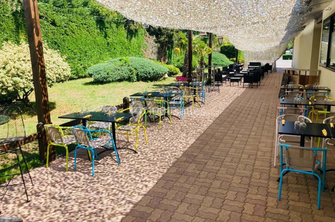 Terrasse extérieure avec tables et chaises colorées, ombragée par un filet, entourée de verdure.