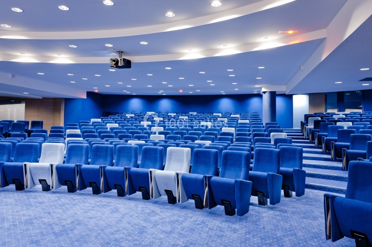 Salle de conférence moderne avec des rangées de sièges bleus vides et un éclairage au plafond.